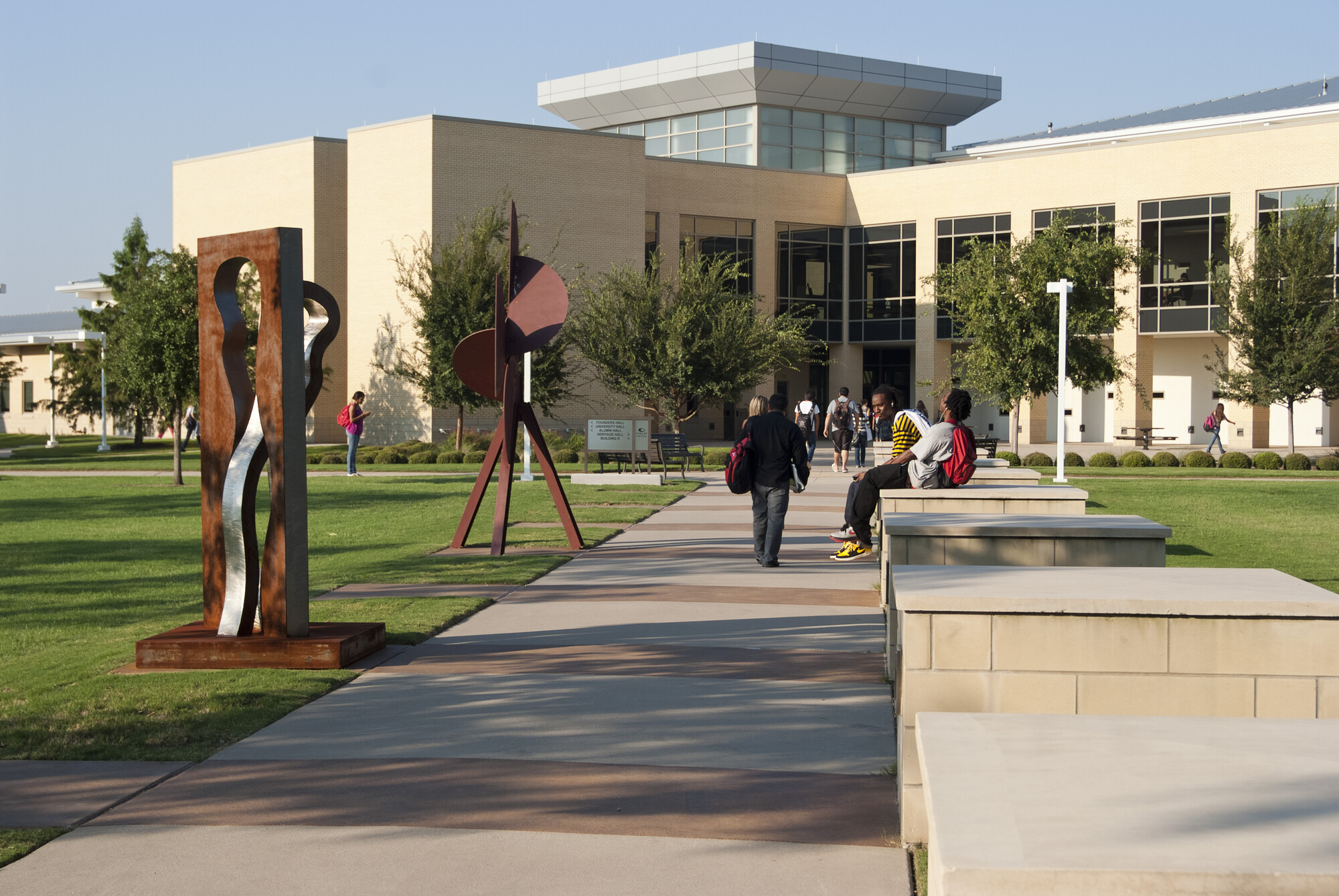 An outdoor photo of Collin College. Students walk down the sidewalk towards the main building. Two tall abstract art sculptures stand in the grass.