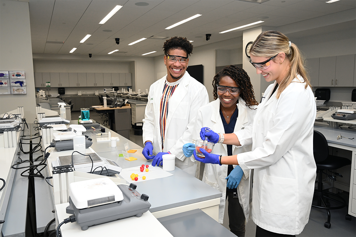 Full uncropped photo of three UNT Health Fort Worth College of Pharmacy students in white lab coats and blue protective gloves work together in a pharmacy skills laboratory.