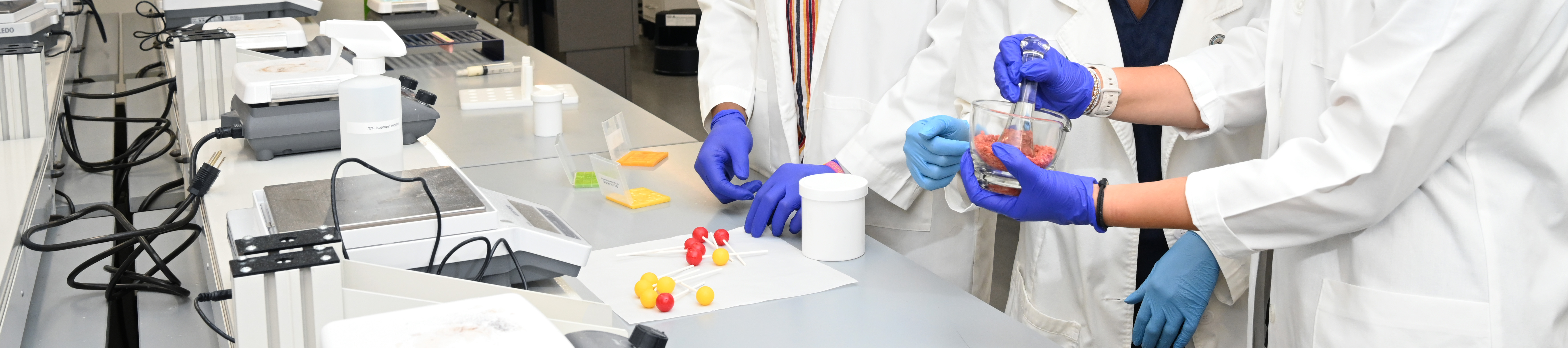 Top vertically cropped photo banner of Full Photo of three UNT Health Fort Worth College of Pharmacy students in white lab coats and blue protective gloves work together in a modern pharmacy skills laboratory, smiling while conducting a hands-on experiment with colorful capsules and compounds.