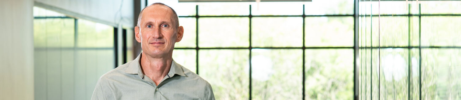 Top wide-narrow cropped banner photo of Andrey Voevodin, Professor of Materials Science and Engineering and Associate Dean for Research at the University of North Texas College of Engineering, stands in a modern academic building lobby wearing a light green button-down shirt.