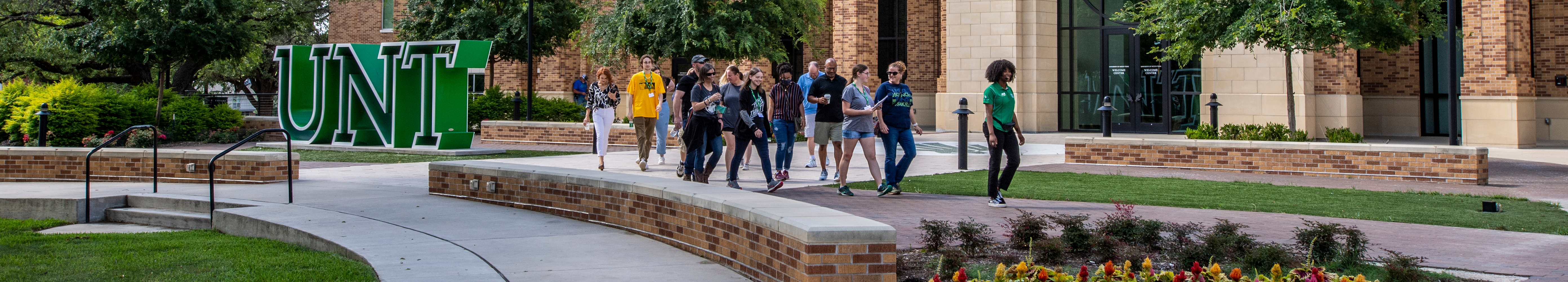 Top horizontal photo banner of UNT's Welcome Center with a very large green and white inlined letters making the UNT logo on the grass in front. A tour of prospective students with their parents that are lead by a official UNT student tour guide are walking by.