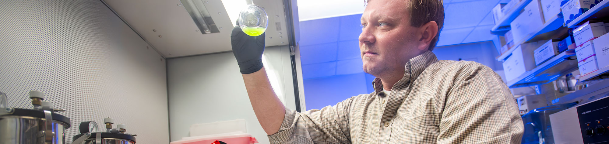 Top horizontal photo banner of UNT's Calvin Henard looking at the bottom of a glass beaker with some lime green liquid that he is holding up to examine in his lab