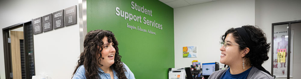 Top horizontal photo banner of two students talking together at a wide table at the UNT Student Support Services office, with one using a laptop.