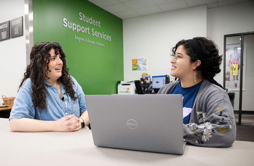 The full photo of 2 UNT students are having a friendly conversation at a table in a UNT Student Support Services office, with one using a laptop.
