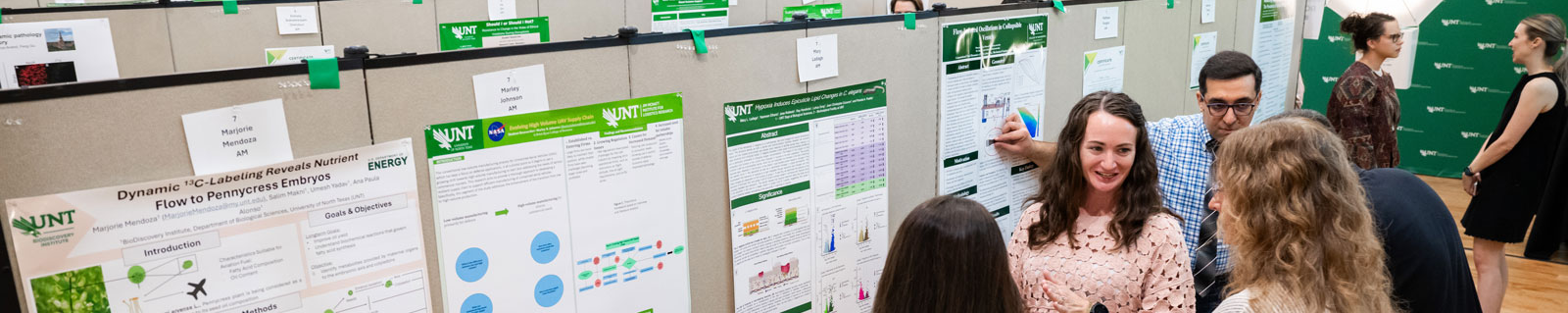Top horizontal narrow cropped photo banner of students presenting their research posters to attendees at the UNT University Research Day in a large indoor hall.