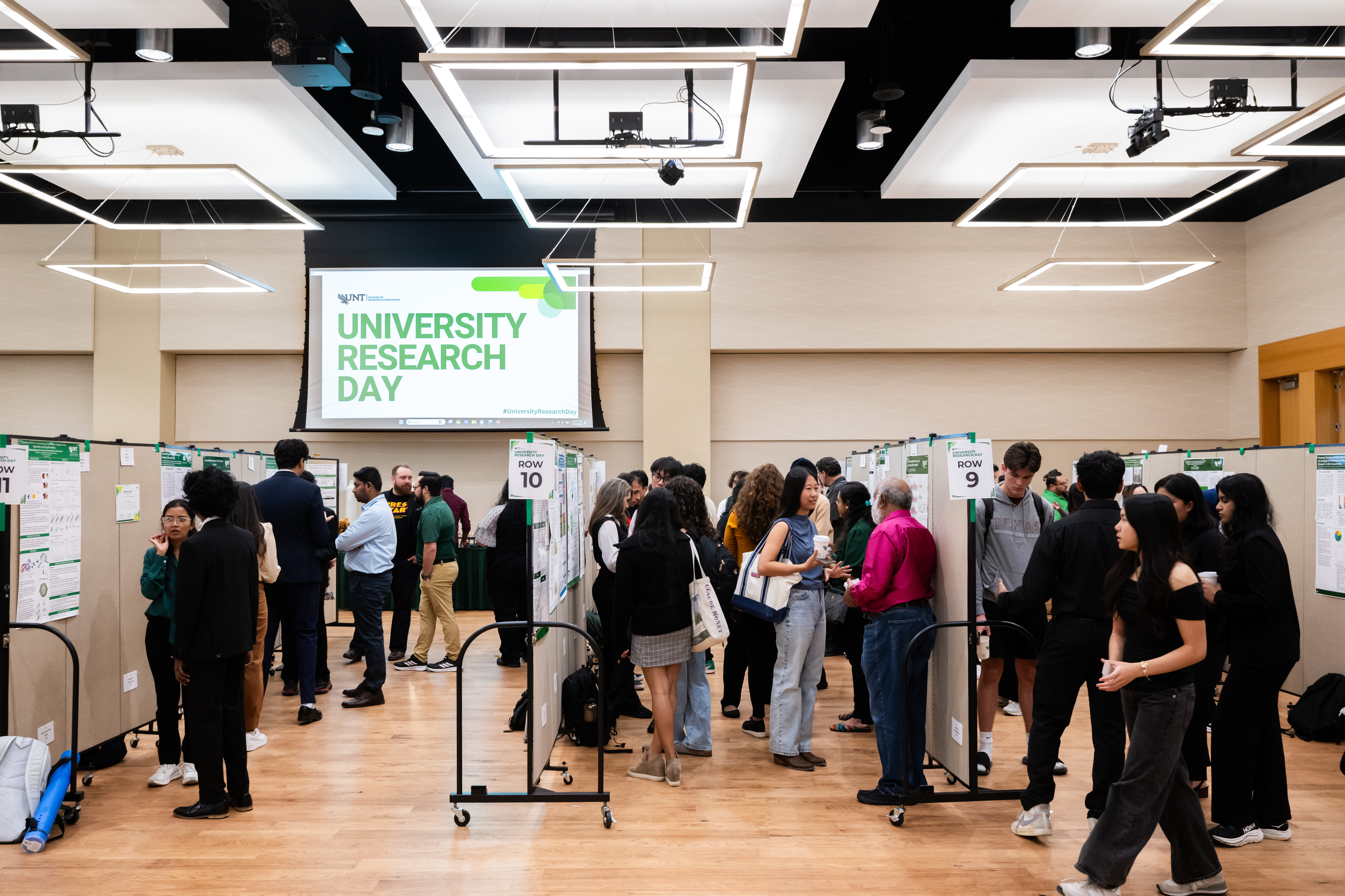 A photo of the crowded exhibit room with UNT attendees exploring the rows of displays at the morning Poster Session hanging on moveable wheeled wall panels.