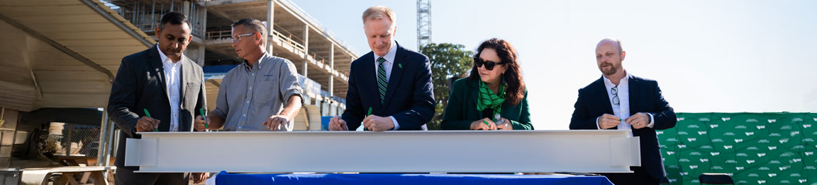 Top horizontal narrow cropped photo banner shows a group of five people signs a steel beam at a construction site with a crane in the background. Standing in the middle is UNT president Dr. Harrison Keller.