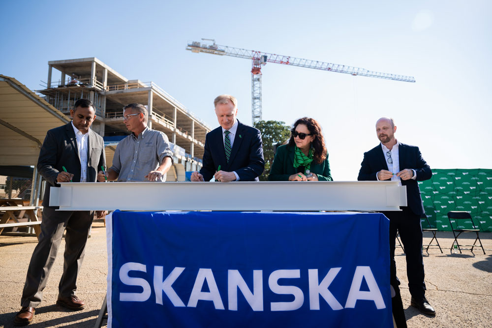 Full photo of a group of five people signs a steel beam at a construction site with a crane in the background. Standing in the middle is UNT president Dr. Harrison Keller.