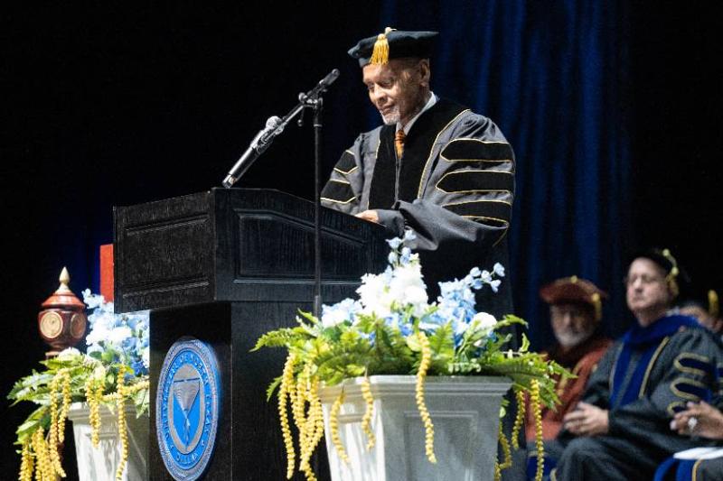 Roland Glynn Parrish in doctoral academic regalia addresses the audience from a flower-adorned podium bearing the UNT Dallas university seal. Photo of Roland Glynn Parrish in academic regalia — a black doctoral gown with gold trim and a mortarboard cap — speaks at a podium during a graduation ceremony. The dark wooden lectern bears a university seal. The stage is decorated with floral arrangements of blue and white flowers with trailing yellow greenery. Two other individuals in academic regalia are seated in the background.