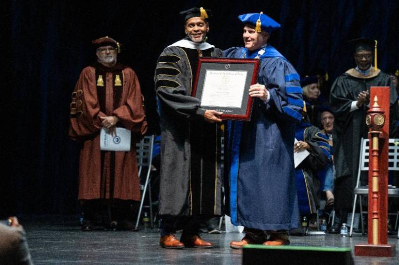 Roland Parrish and UNT Dallas President Warren von Eschenbach hold a framed honorary Doctor of Humane Letters degree certificate together on stage during the Spring 2026 Commencement ceremony. Roland Parrish and UNT Dallas President Warren von Eschenbach in academic regalia stand center stage holding a framed honorary degree certificate during a university commencement ceremony. One wears a black doctoral gown with gold trim; the other wears a blue doctoral gown. Additional faculty members in academic regalia are visible in the background, with one applauding.