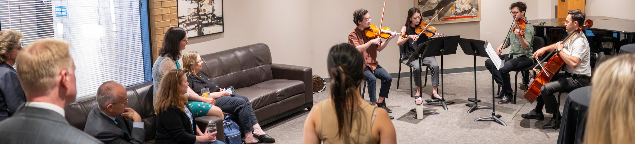 Top horizontal narrow cropped photo banner of four University of North Texas College of Music students perform a chamber string quartet — two violinists, a violist, and a cellist — reading from music stands before a small seated audience in an a warmly lit reception room featuring abstract paintings, Denton, Texas. 