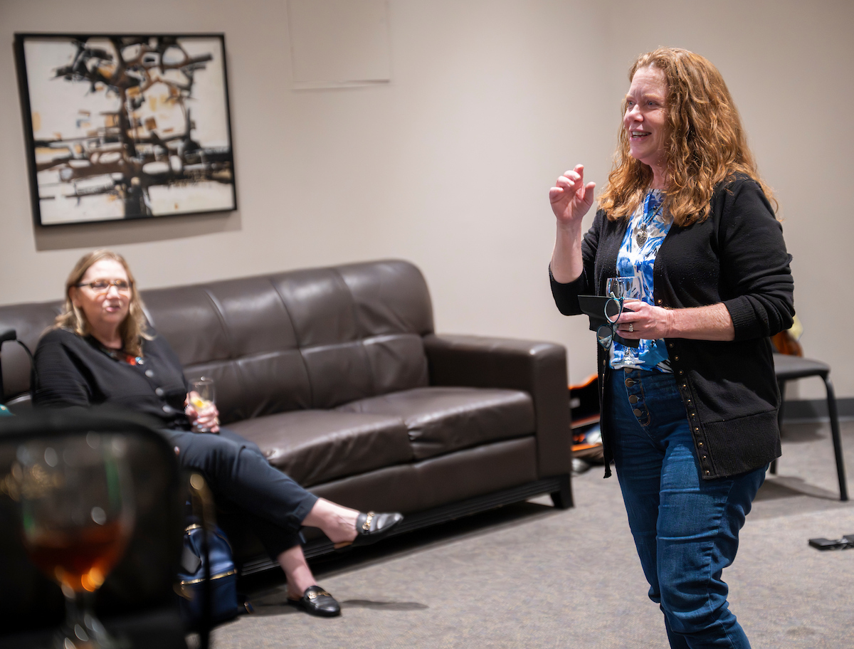 Photo of Deborah Watkins, UNT alumna and donor, speaks to guests while holding a wine glass at an intimate reception in the UNT College of Music, with abstract artwork visible on the wall behind her.