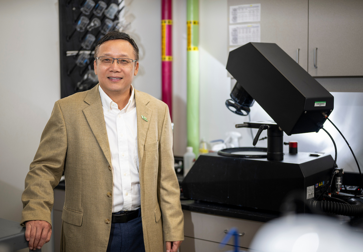UNT's Jincheng Du stands beside advanced research equipment inside a university science laboratory. A portrait photo of UNT faculty member Jincheng Du wearing a tan blazer and UNT lapel pin stands beside advanced research equipment in his Functional Glasses and Materials Modeling Laboratory at the University of North Texas.