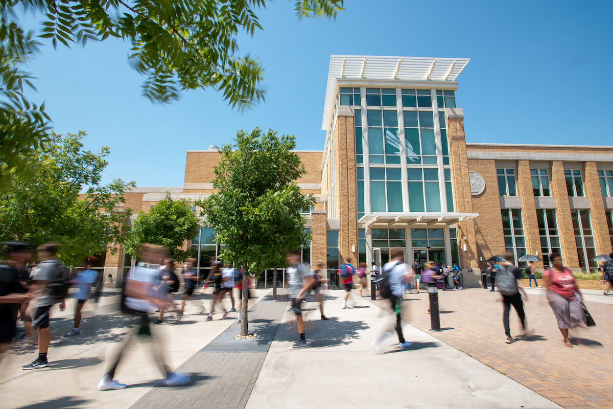 Photo of UNT motion blurred students walking in front of the Student Union, a modern brick and glass campus building on a sunny day.