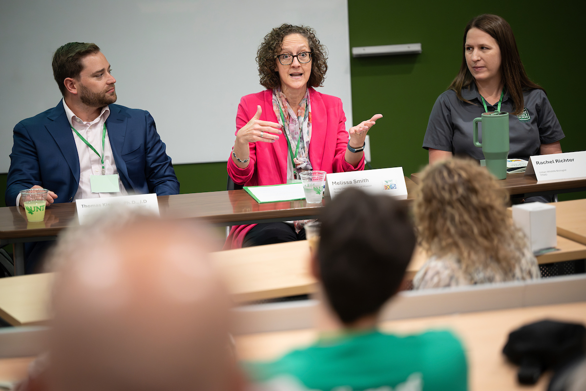 Photo of a group of onlookers look at the three panelists, one man and two women, the woman in the center is talking