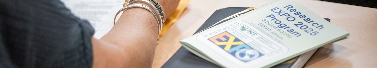 Top horizontal photo banner of a closed program guide with the title of Research Expo 2025 program positioned on top of a closed black notebook to the right of a student's arm resting on a table.