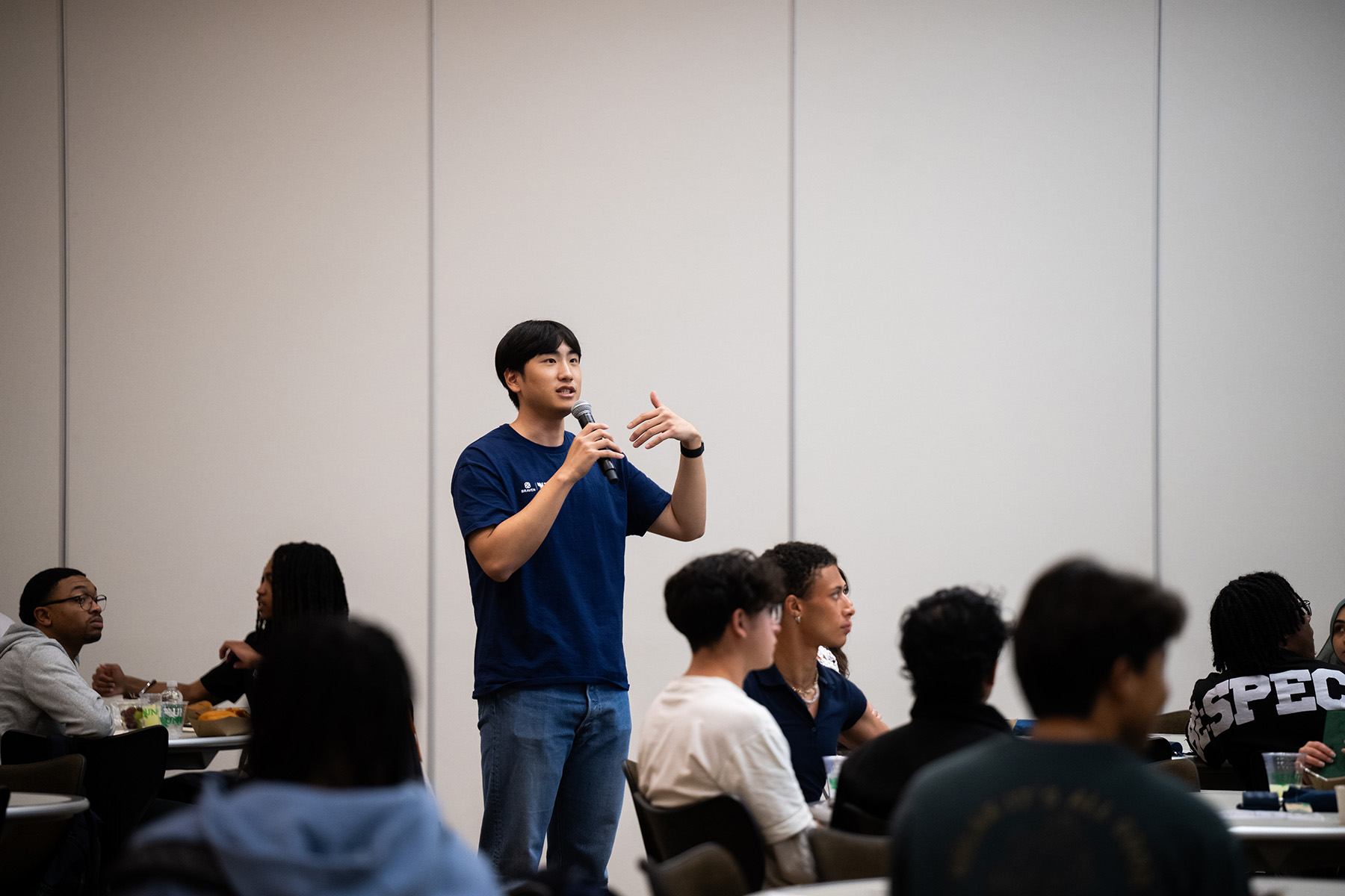 William Kim, facilitator for the UNT Braven Career Accelerator, addresses students at Live Your Legacy Photo of William Kim, a person wearing a navy shirt stands and speaks into a microphone while people seated at tables listen, who is a facilitator for the UNT Braven Career Accelerator, addresses students at Live Your Legacy