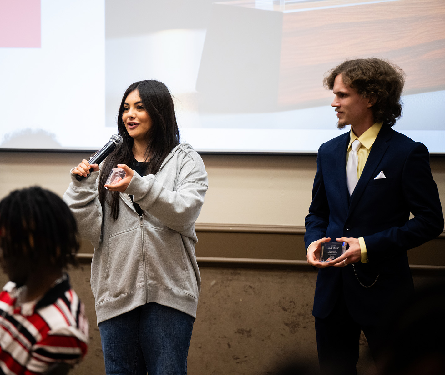 UNT's Madeline Ledesma (left) accepts a Live Your Legacy award Photo of UNT's Madeline Ledesma (left), A young woman in a gray sweatshirt speaks into a microphone while a young man standing to her right listens, while she accepts her Live Your Legacy award