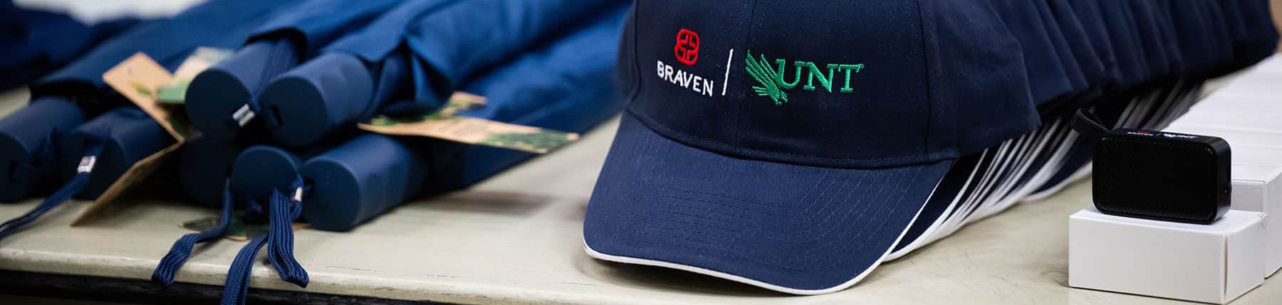 Top horizontal narrow cropped photo banner of navy baseball cap embroidered with Braven and UNT logos displayed on a table alongside navy rolled umbrellas at a UNT Braven Career Accelerator event.