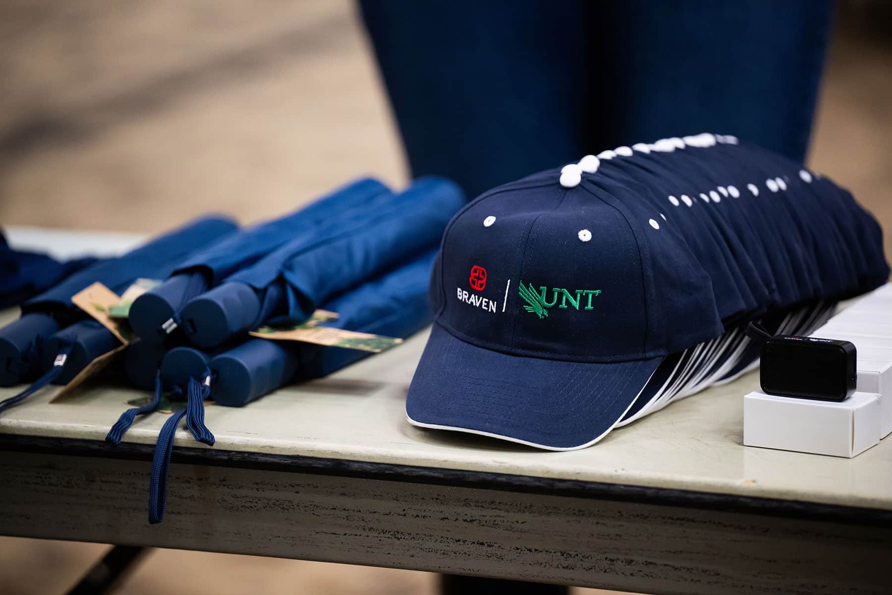 Full uncropped photo of a navy baseball cap embroidered with Braven and UNT logos displayed on a table alongside navy rolled umbrellas at a UNT Braven Career Accelerator event.