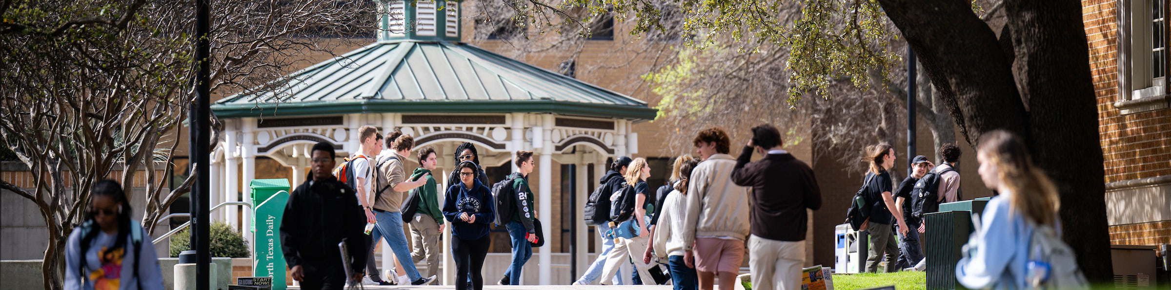 Top horizontal, narrow-cropped photo banner of a wide view of a UNT students walking across a busy university campus near a gazebo.