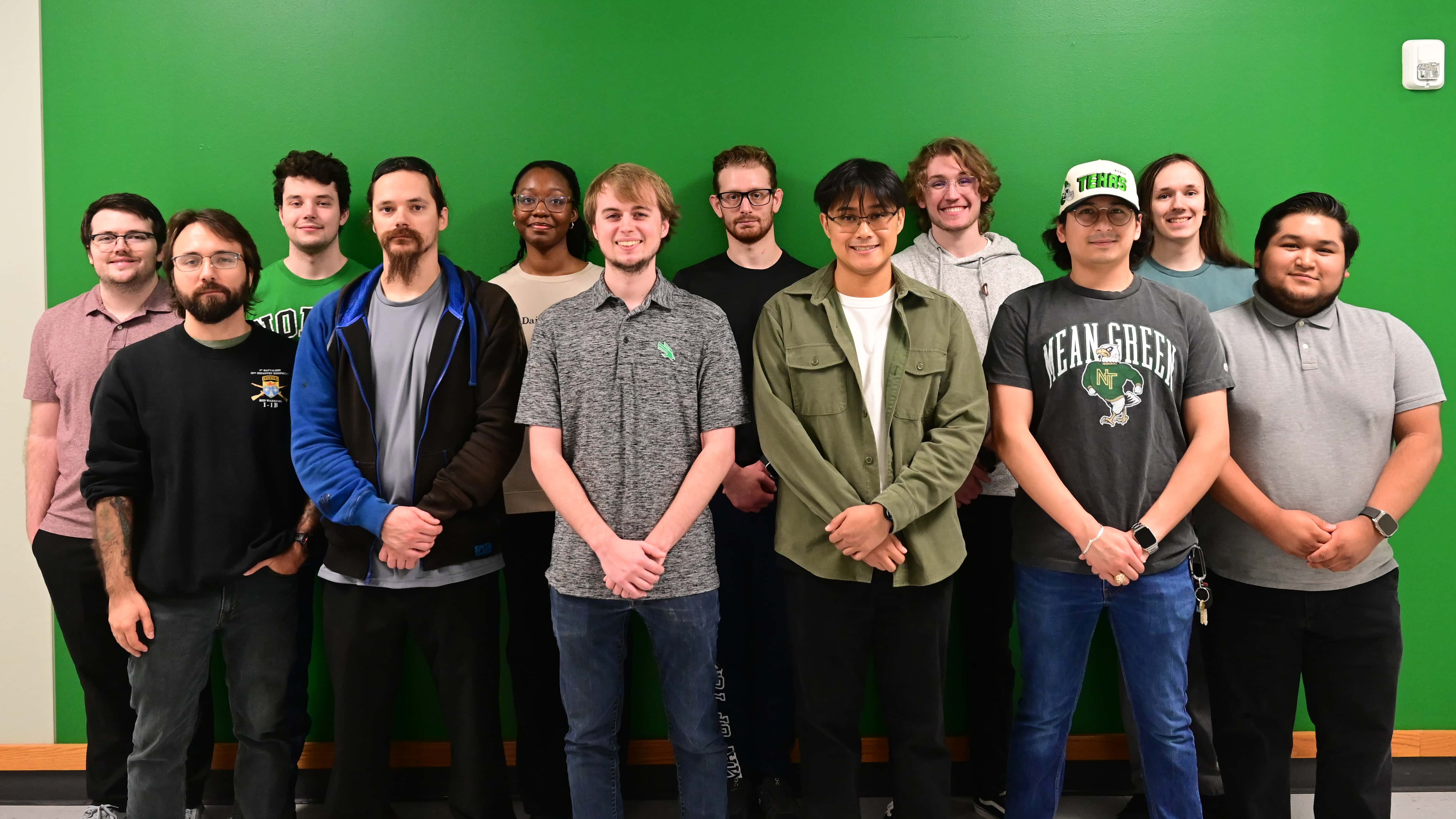 A group of 12 UNT students stand in front of a green wall, six in the front, six in the back. Many are smiling at the camera.