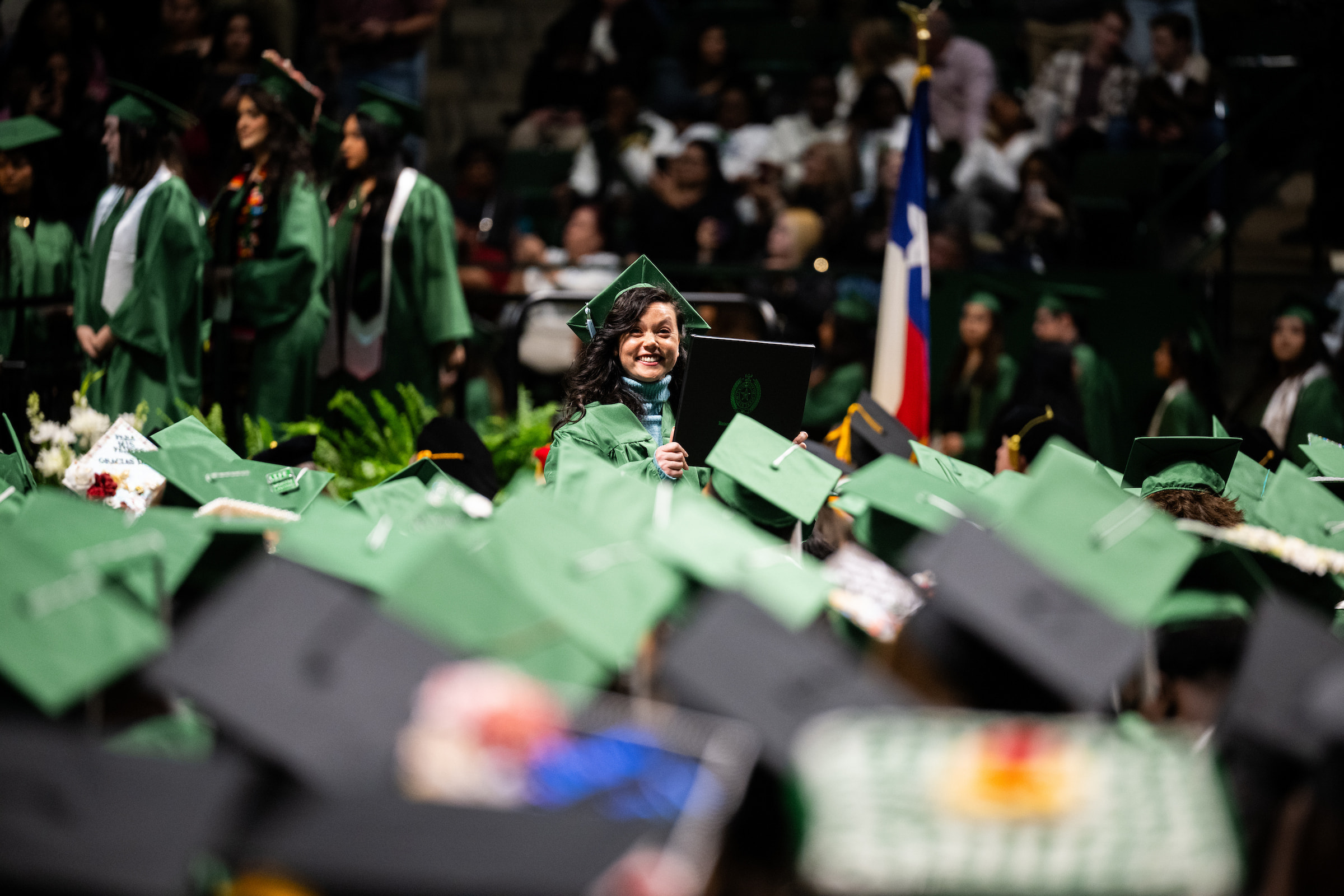 Full photo of a smiling new UNT graduate in green cap and gown holds up her diploma amid rows of fellow graduates during the University of North Texas Commencement ceremony, with the Texas state flag visible in the background.