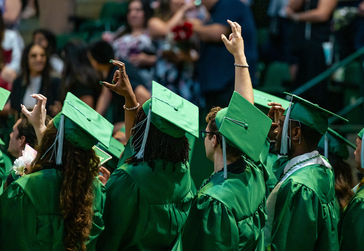 Photo of UNT graduating students wearing their green cap and gown raising their hand giving the school's eagle claw salute 