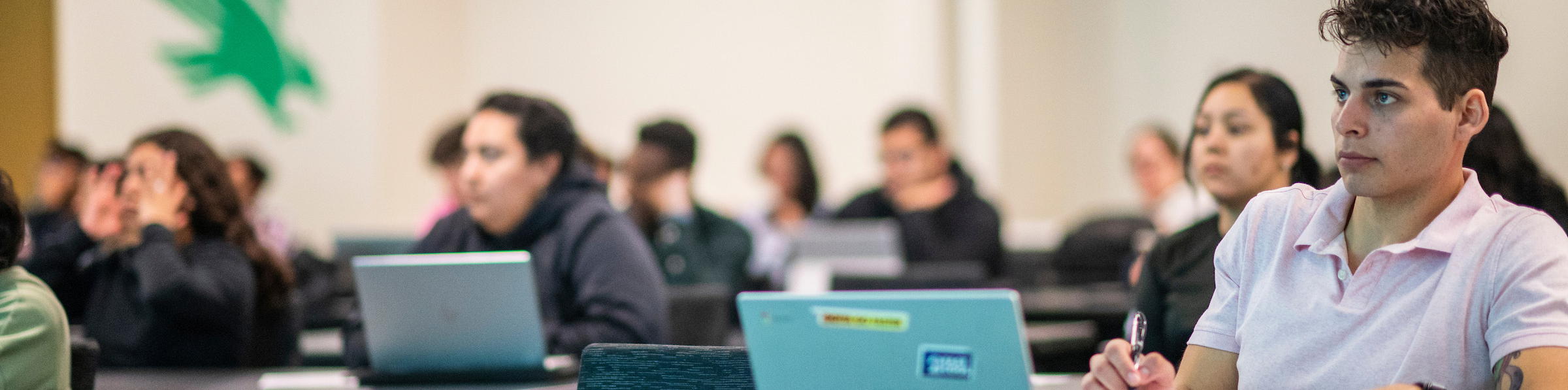 Photo of top wide-narrow cropped banner photo of selective focus on UNT students with laptops attentively listening in a classroom, with the UNT Mean Green Eagle logo on the wall in the back slightly out of focus.