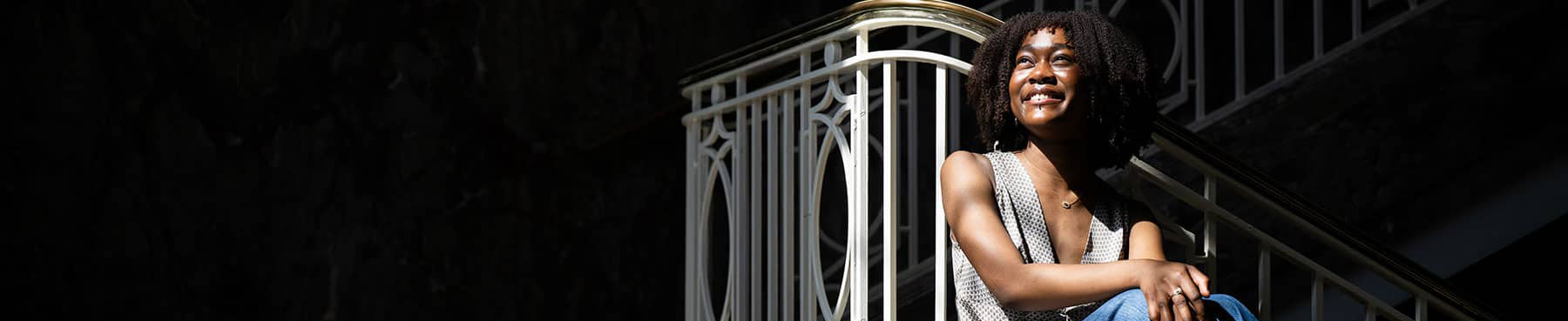 Top wide-narrow cropped banner photo of a smiling Hadiyah "Diyah" Muhammad, UNT Honors College international studies senior and Rotary Global Grant recipient, seated on floor, leaning against an art deco ornate white staircase railing with marble steps. The photograph has strong dynamic shadows that fill most of the composition with some highlights that illuminate Diyah.