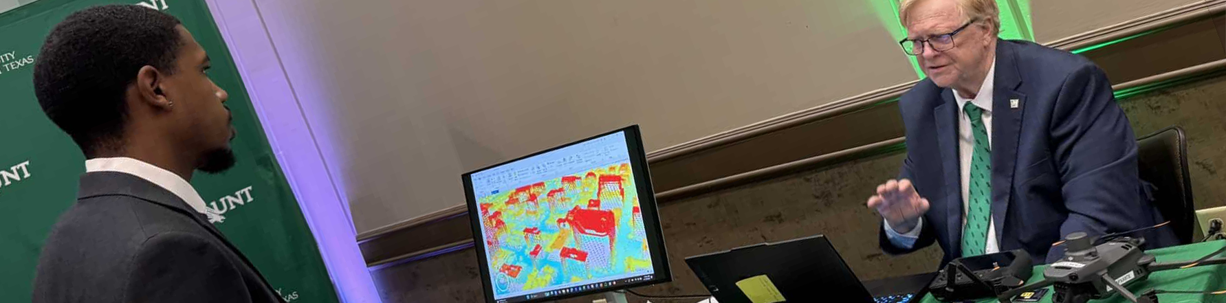 Top wide-narrow banner photo of a man standing in front of a table of books and a computer monitor, behind which another man is seated. He is the UNT Geography & the Environment lecturer, John South, providing information on drones.