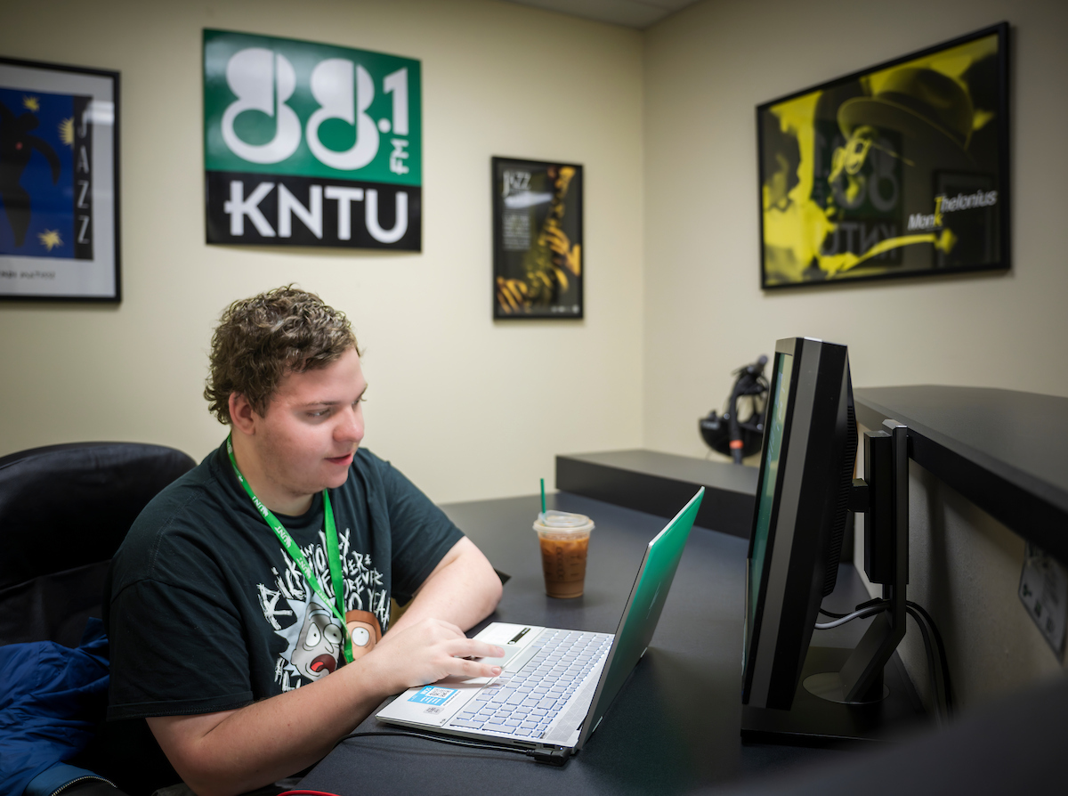 Photo of UNT's Alex Bartolo sitting at a desk working on a laptop in the KNTU radio station office
