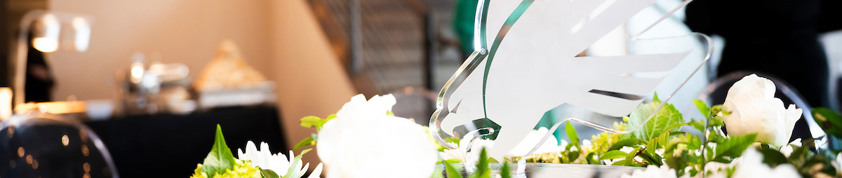 Top photo banner from UNT's Salute to Faculty Excellence awards celebration at the venue. The picture is a close up of a table top covered with white flowers and a variety of greenery at the base of a crystal sculpture of the diving eagle logo symbol.