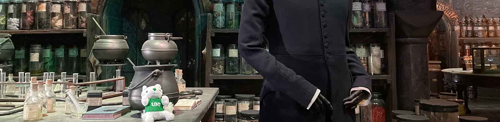 This top cropped banner image shows a detailed potion classroom setup from the Harry Potter movie series, with cauldrons, glass bottles, herbs, and a mannequin in dark robes, alongside a small plush white squarel toy wearing a green "UNT" shirt.