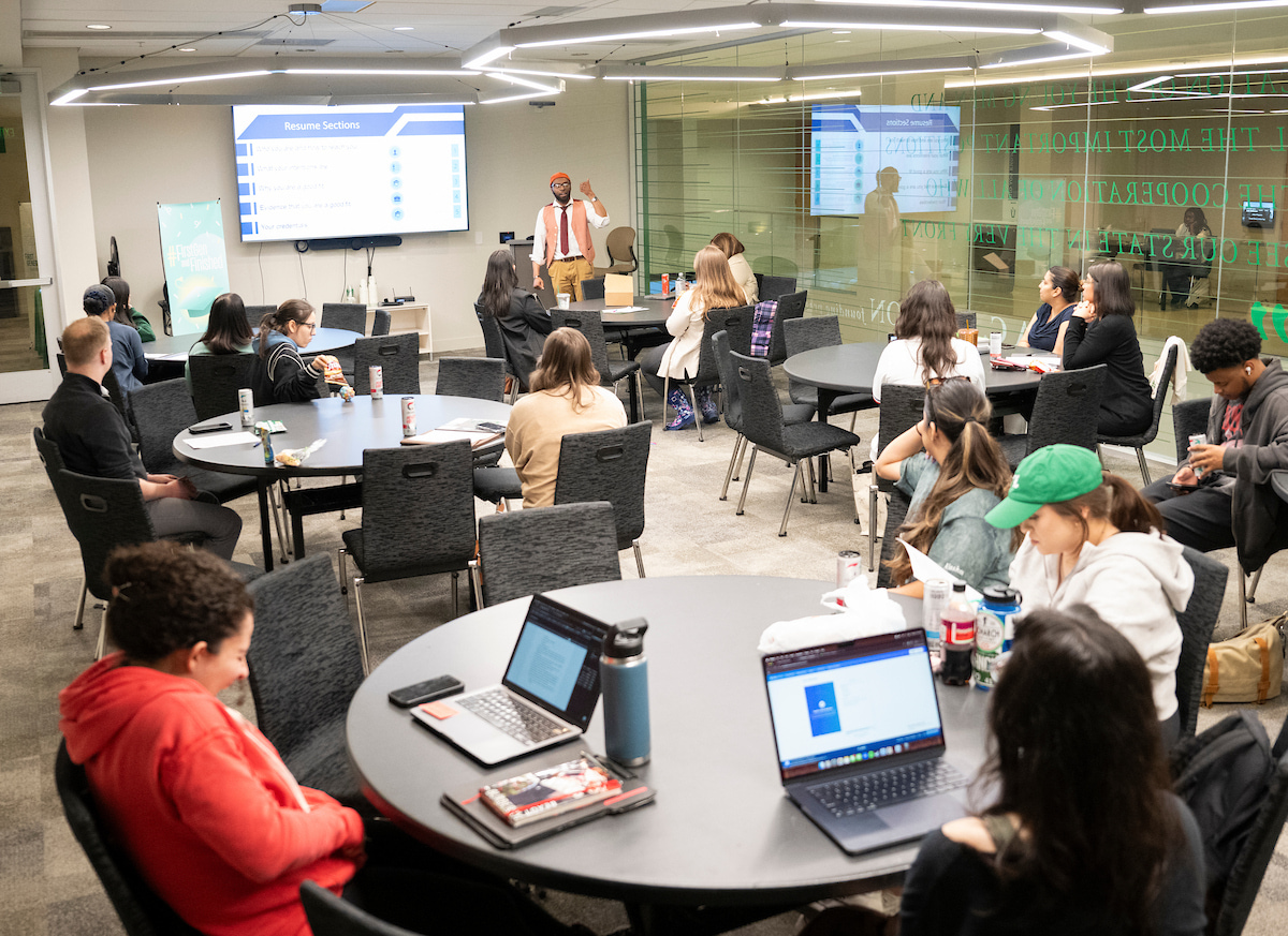 A presenter leading a workshop for UNT students seated at round tables with laptops. Photo of a presenter leading a workshop for UNT students seated at round tables with laptops.