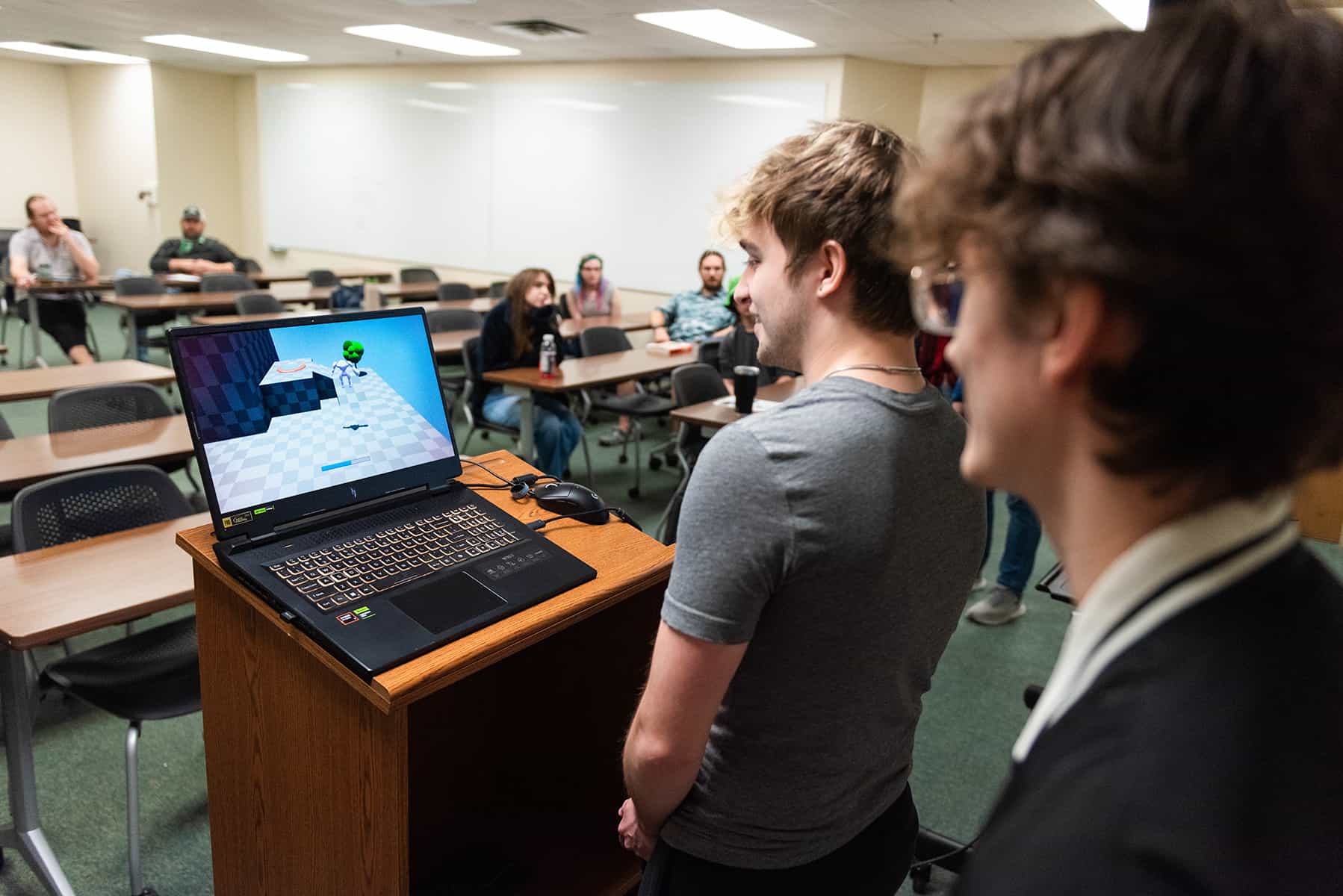 Photo of UNT students in a college classroom with two standing in the foreground by a lecture podium with an open laptop on it. The illuminated screen shows a digital 3D game graphic. The angle of the perspective is looking back into the room