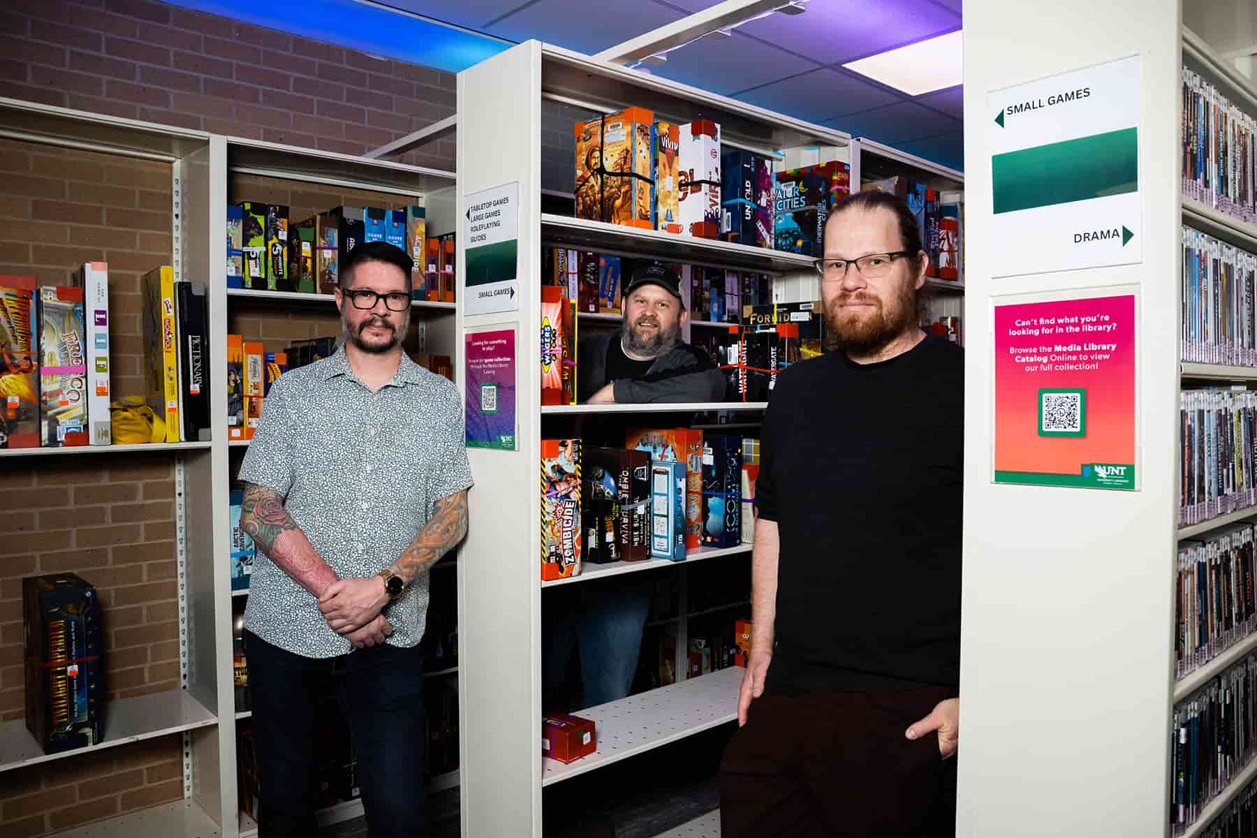 Photo of 3 men standing by a library with stacks (floor to ceiling rack of shelfs) holding digital games in their retail boxes. Starting from the left side are UNT's Darin Bradley, assistant professor of practice in English; Gabriel Olson, program director and professor of practice in video games; Stephen Mandiberg, senior lecturer in media industries and video games