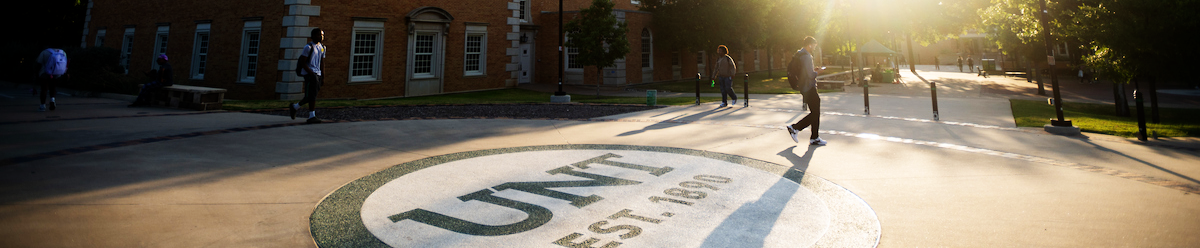 Top horizontal narrow cropped photo banner shows The University of North Texas Hurley Administration Building with the sun setting behind it, with a few students walking nearby. 