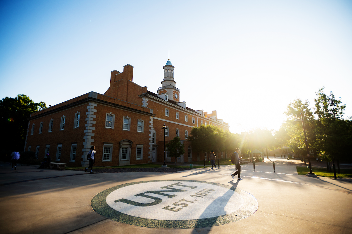 Full photo of The University of North Texas Hurley Administration Building with the sun setting behind it, with a few students walking nearby. 