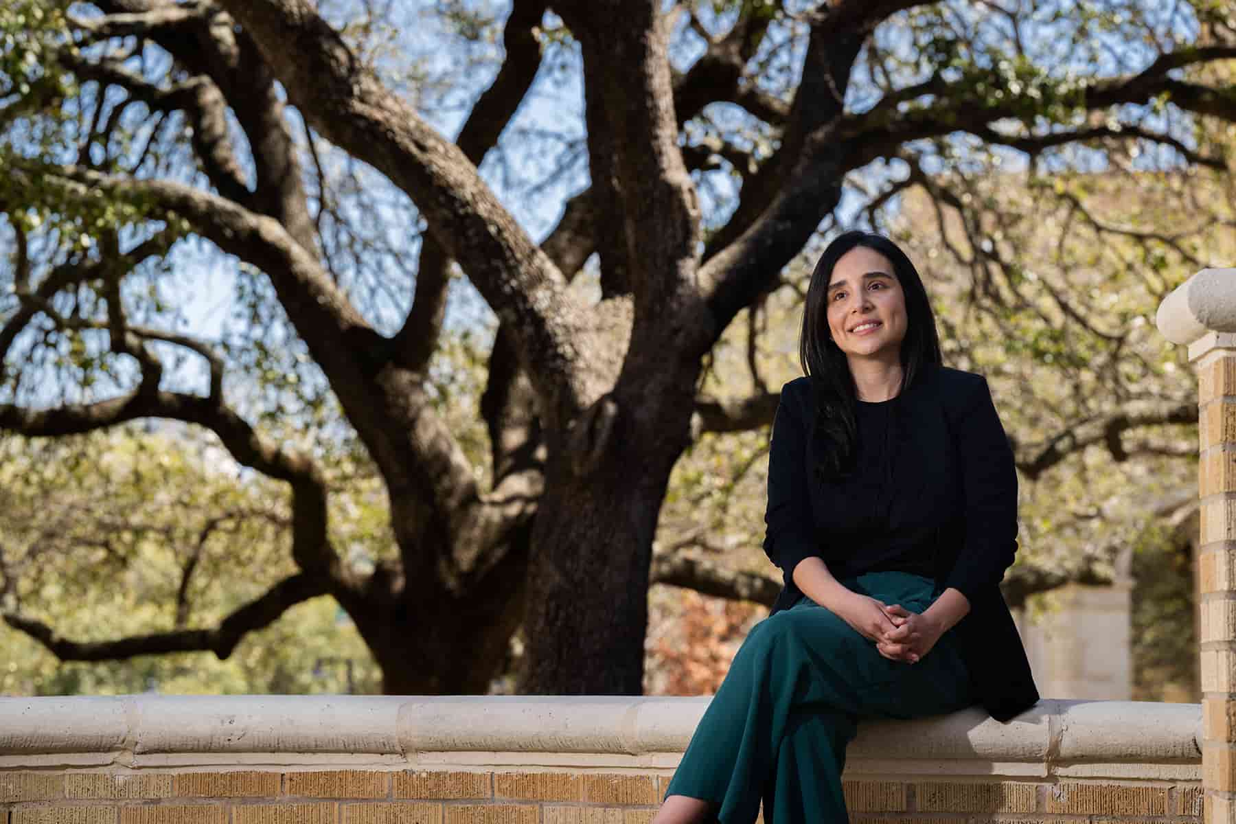 Full photo of Isamar Almeida, UNT behavioral science Ph.D. graduate, seated outdoors on campus with a large oak tree in the background.