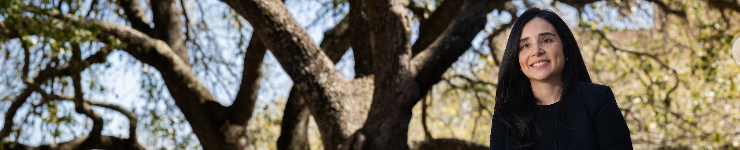 Top wide-narrow cropped banner photo of Isamar Almeida, UNT behavioral science Ph.D. graduate, seated outdoors on campus with a large oak tree in the background.