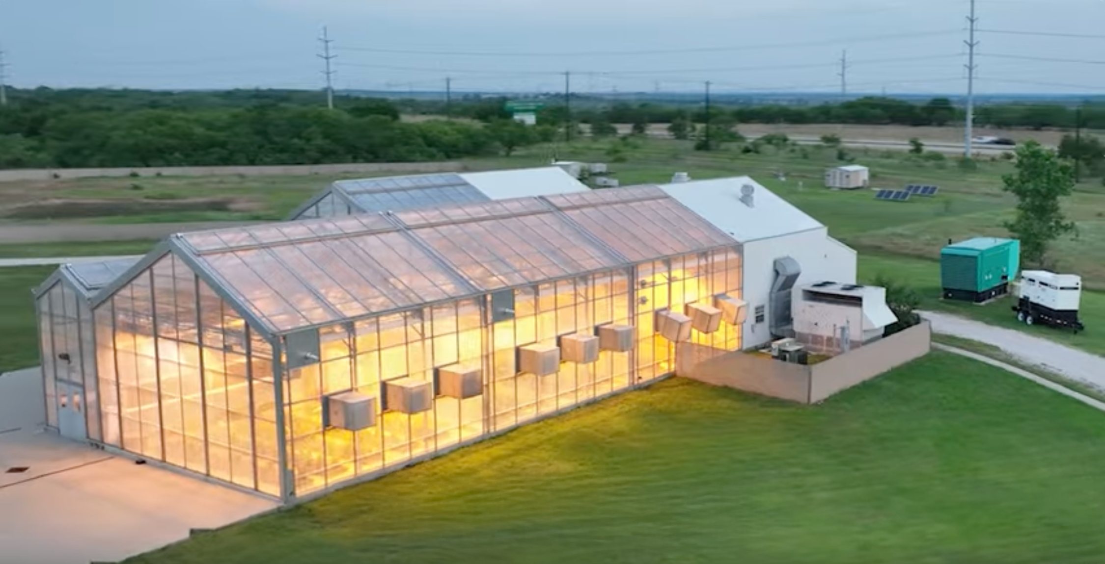 A photo of an illuminated the UNT Discovery Park Campus Greenhouse is shown from the outside during dusk, surrounded by grassy fields and equipment.
