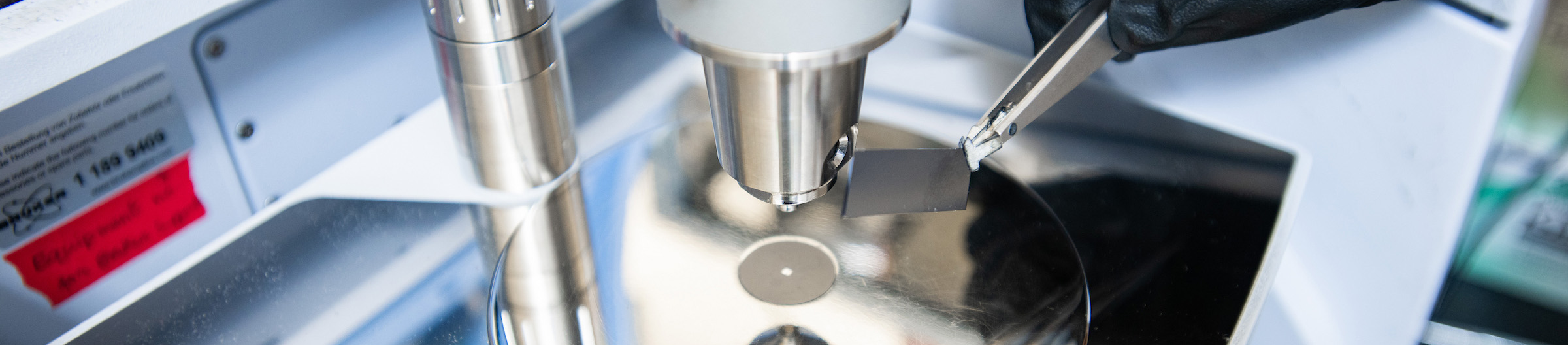 Top horizontal photo banner of a close-up at a UNT research laboratory setting. A gloved hand uses tweezers to position a small, dark square material beneath a spectrometer, near the center of the platform.
