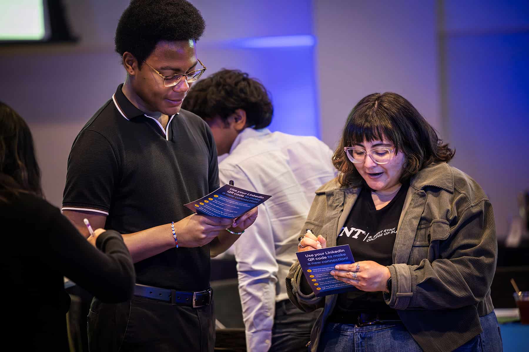 Two UNT students stand together reviewing LinkedIn-branded pamphlets titled 'Use your LinkedIn QR code to make a new connection' during the Open to Work Lab event, with additional students visible and engaged in activity in the background.