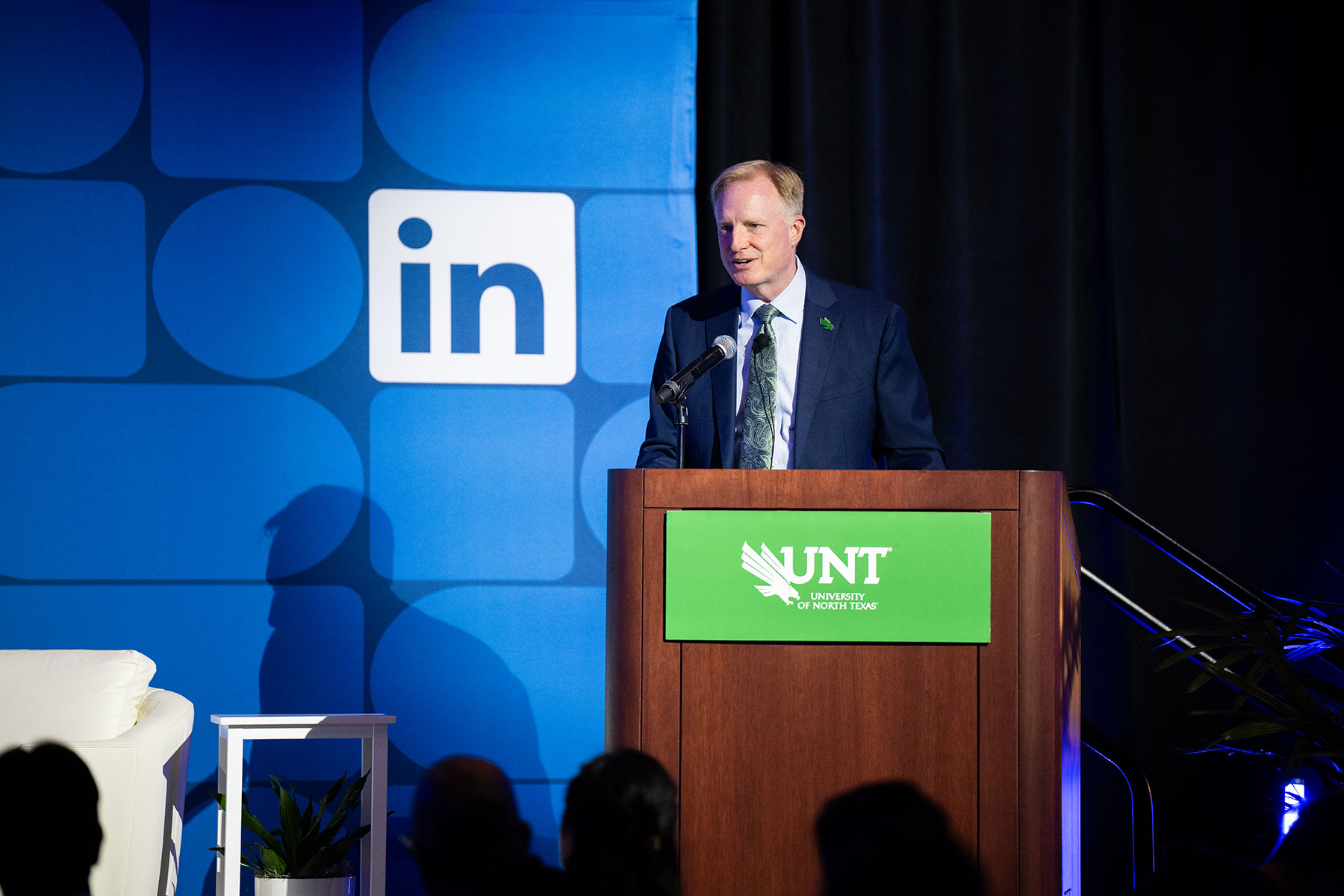 Photo of UNT President Harrison Keller stands at a wood podium bearing the University of North Texas green logo, speaking into a microphone in front of a blue LinkedIn-branded backdrop, with audience members silhouetted in the foreground.