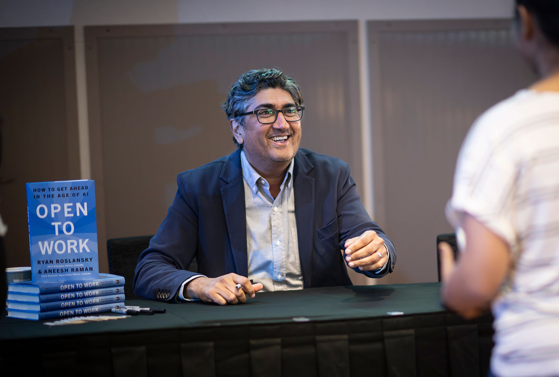 Photo of LinkedIn Chief Economic Opportunity Officer Aneesh Raman sits smiling at a table during a book signing, with a stack of copies of 'Open to Work: How to Get Ahead in the Age of AI,' co-authored by Ryan Roslansky and Aneesh Raman, displayed to his left.