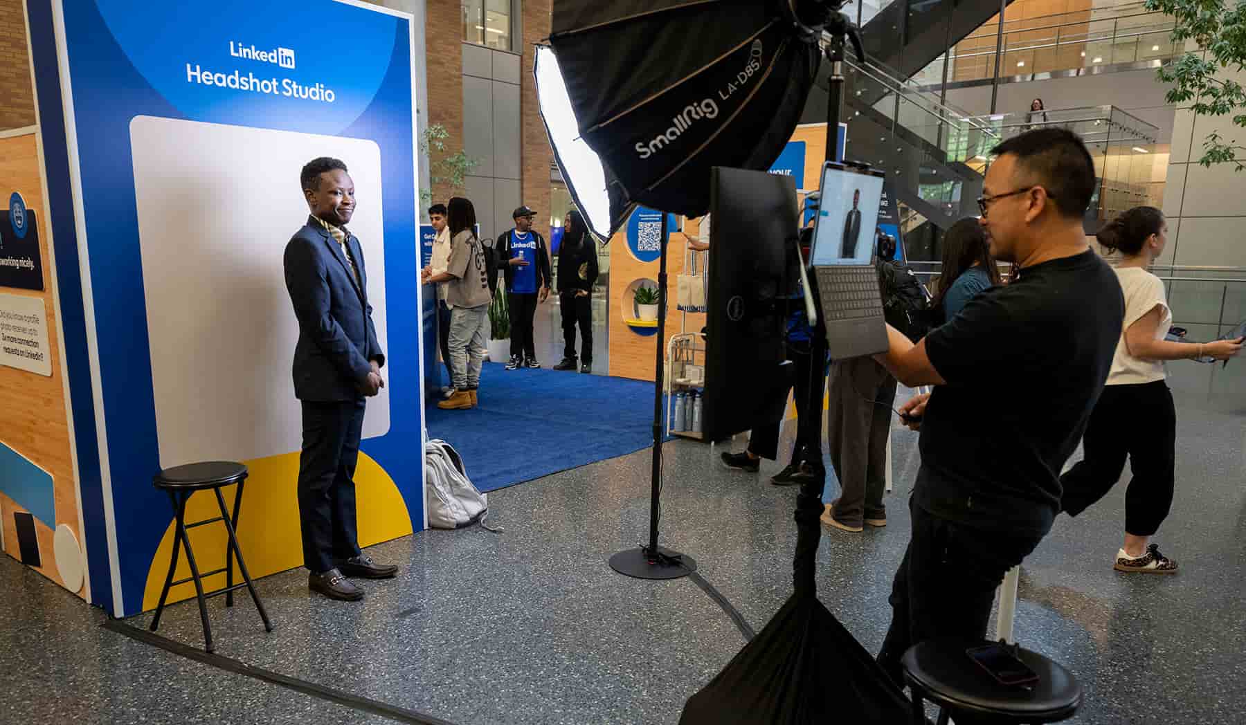 A UNT student in a suit stands in front of the LinkedIn Headshot Studio backdrop while a photographer holds a tablet and professional lighting equipment captures his photo, with other students and event displays visible in the background of the campus building atrium.