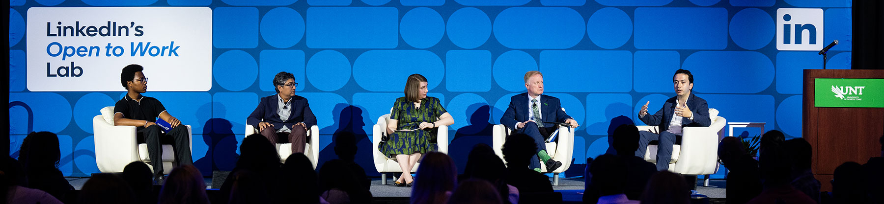 Photo of top wide and narrow cropped banner photo of five panelists seated in white chairs on a stage in front of a blue LinkedIn-branded backdrop reading 'LinkedIn's Open to Work Lab,' with the UNT logo visible on a podium to the right and a full audience seated at tables in the foreground.