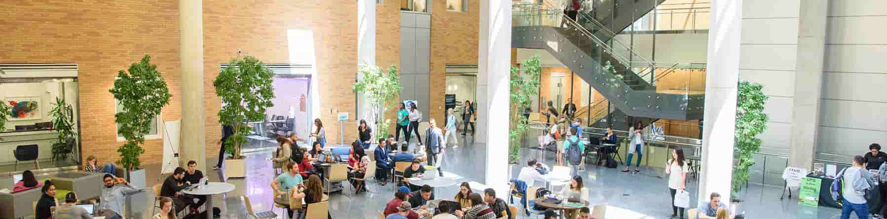 Top wide-narrow cropped banner photo of UNT students studying and collaborating inside the atrium at the Business Leadership Building, Denton Texas with an exposed free standing metal staircase in the background.