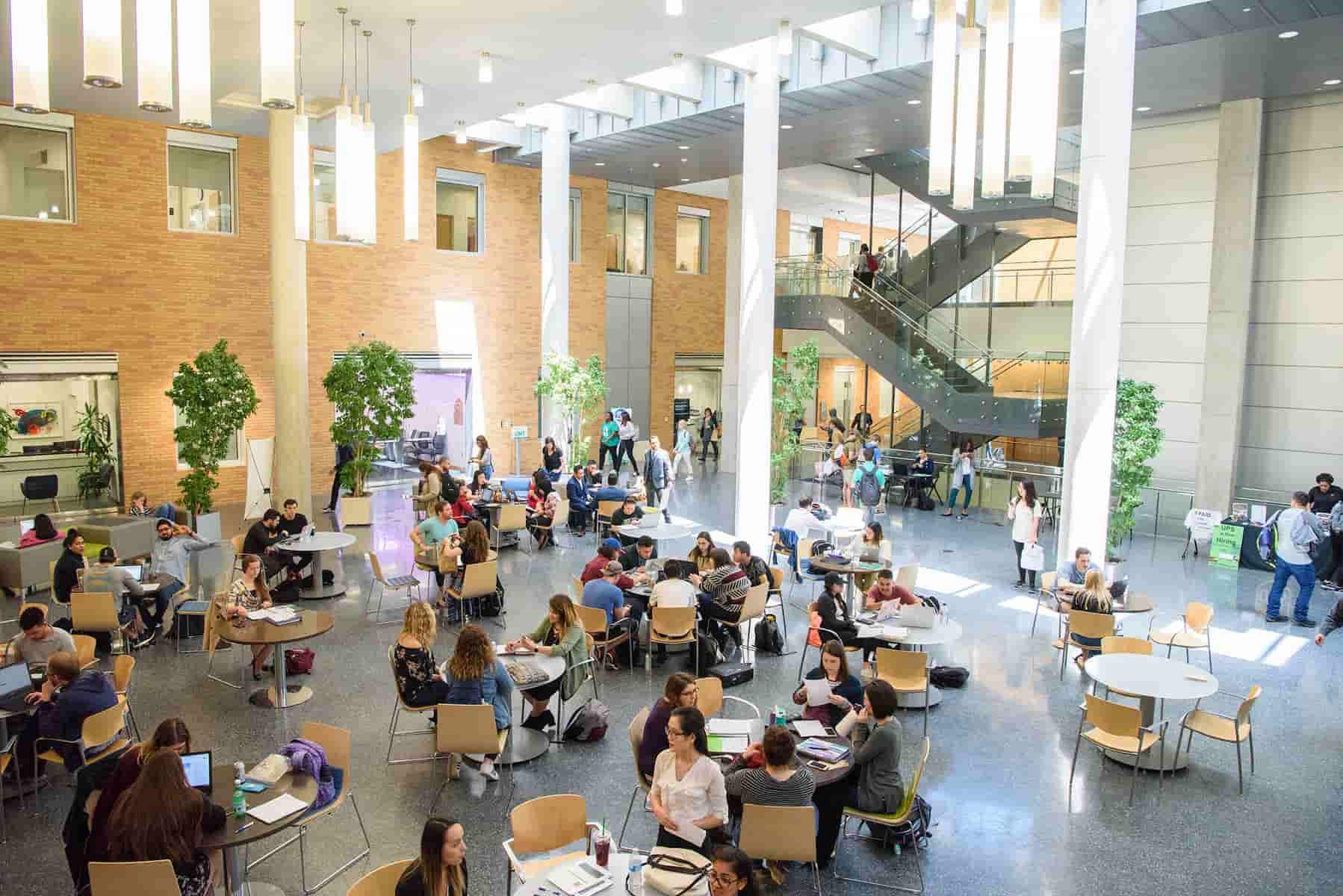 Full photo of of UNT students studying and collaborating inside the atrium at the Business Leadership Building, Denton Texas with an exposed free standing metal staircase in the background. 